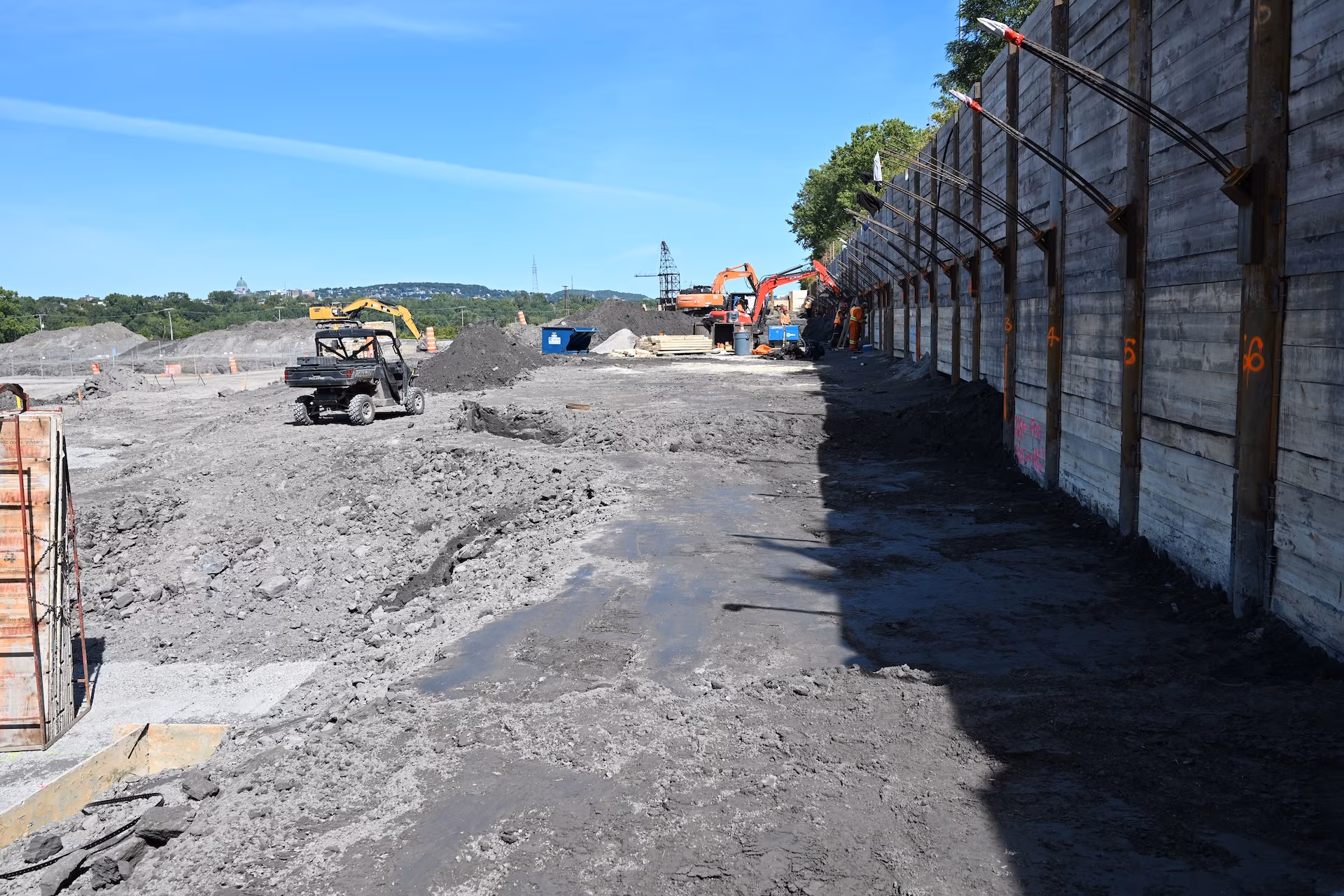 Construction site with heavy machinery, piles of dirt, and workers near a tall retaining wall under clear blue sky.