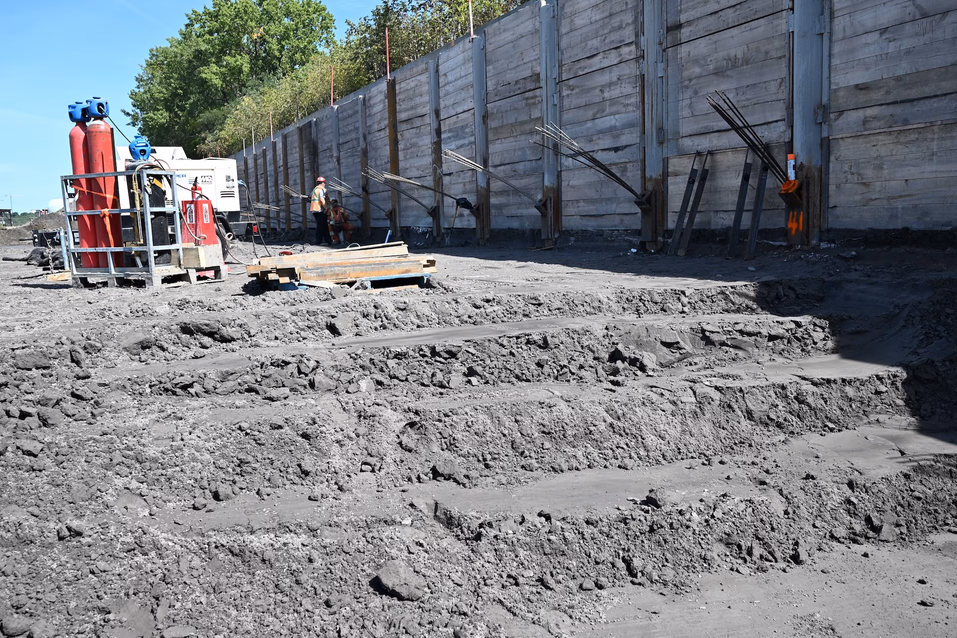 Construction site with layered dirt excavation, two workers near heavy equipment and a tall wooden retaining wall with exposed rebar.