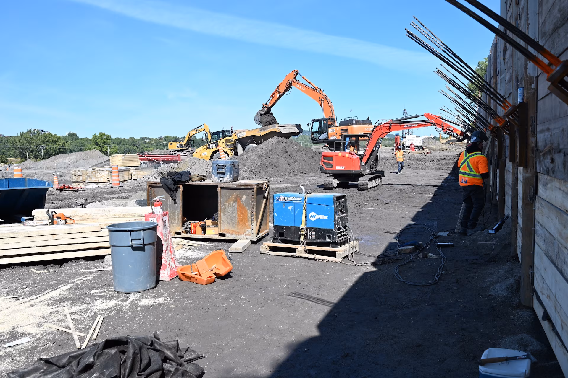 Construction site with workers in safety gear, excavators loading dirt, and various equipment scattered on the ground under a clear blue sky.