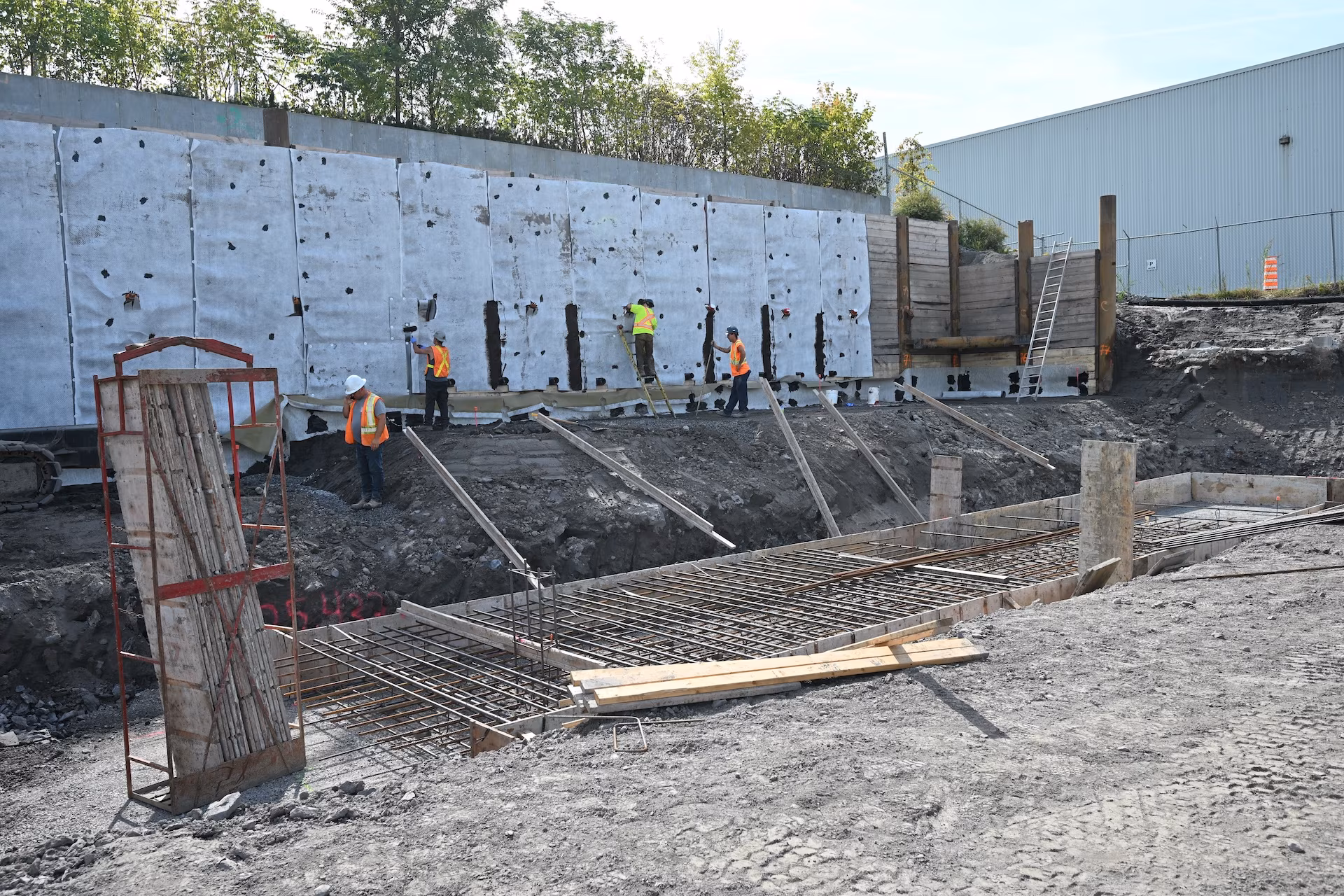 Construction workers in safety gear working on a reinforced concrete foundation near a retaining wall covered with protective sheets.