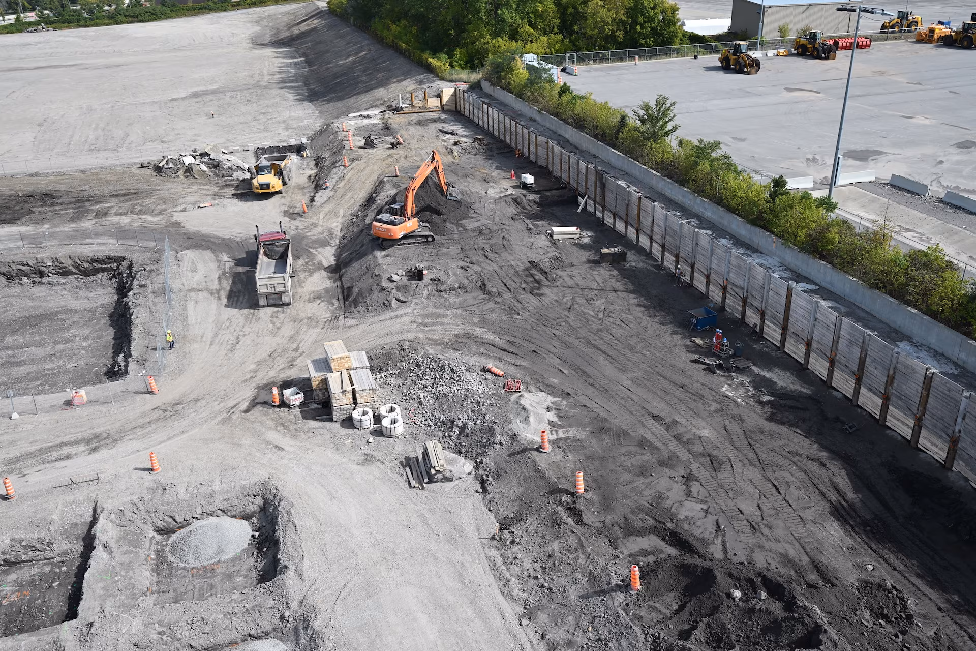 Aerial view of a construction site with an excavator loading dirt into a dump truck, piles of construction materials, and safety cones scattered around.