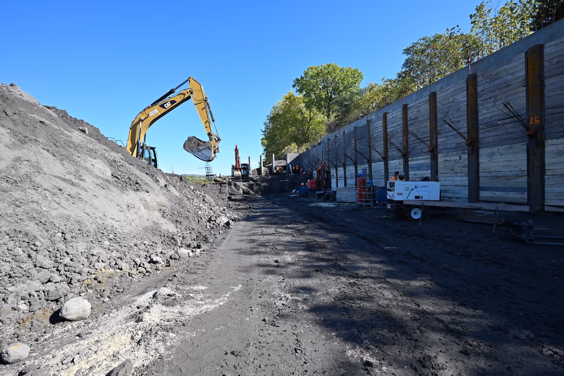Construction site with a yellow excavator moving soil next to a tall reinforced concrete retaining wall under a clear blue sky.