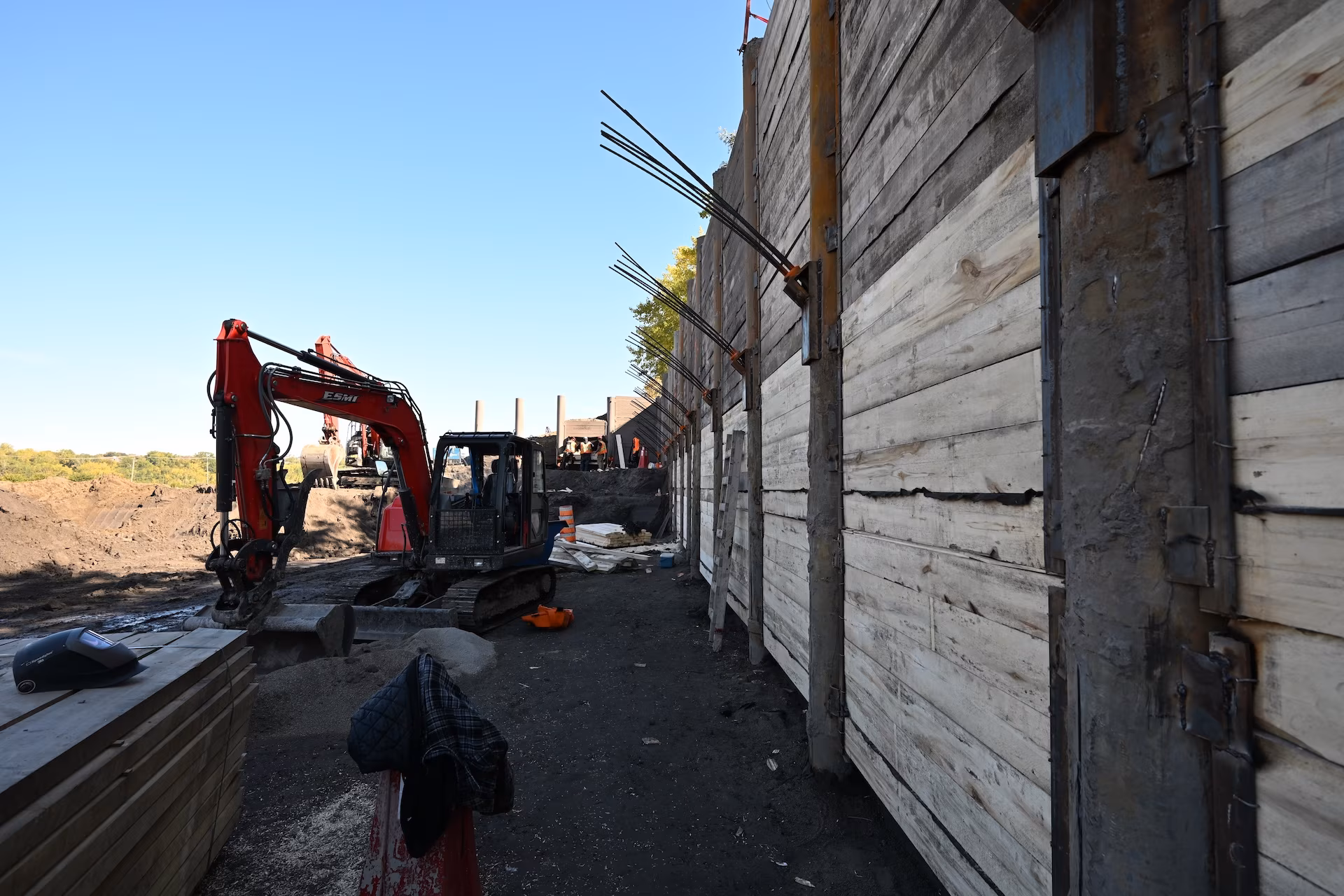 Construction site with a red excavator next to a retaining wall reinforced with wood and metal beams.