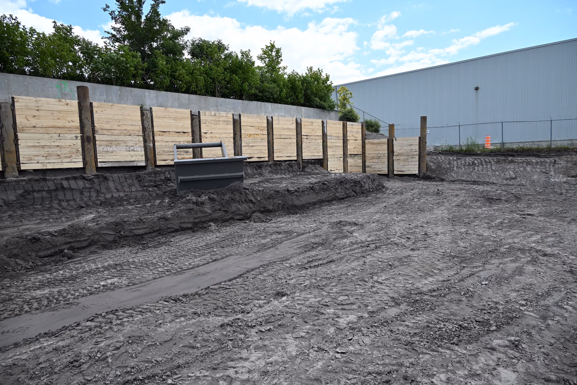 Cleared construction site with a dirt surface, wooden retaining wall supported by vertical posts, and a large industrial building in the background.