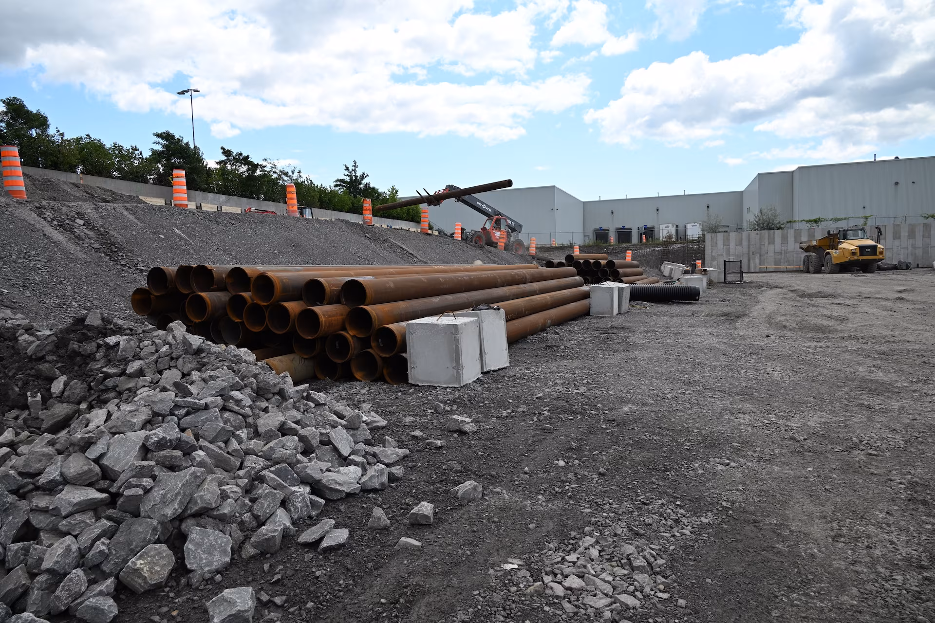Construction site with stacks of large rusty metal pipes, piles of gray rocks, and heavy machinery including a loader and a forklift under a partly cloudy sky.