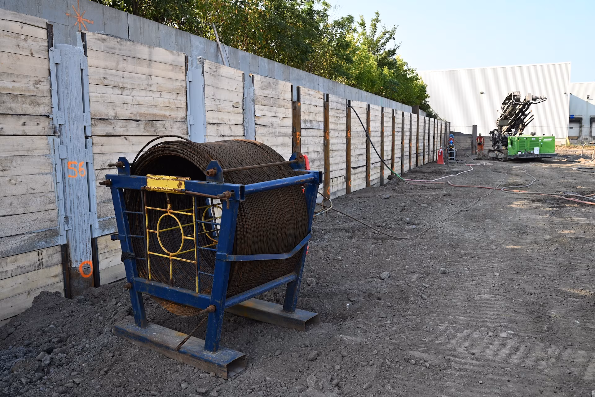 Large spool of coiled steel cable on a blue metal frame at a construction site with wooden retaining walls and machinery in the background.
