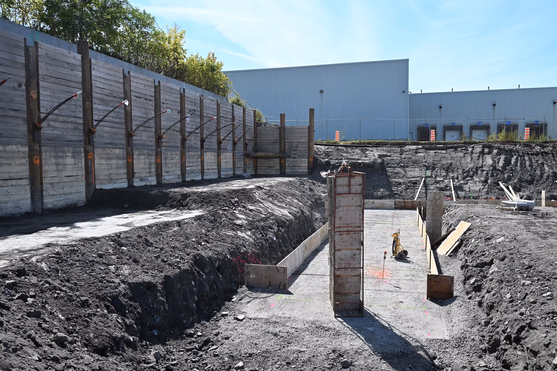 Construction site with deep excavation, wooden retaining wall, and formwork for concrete foundations under a clear blue sky.