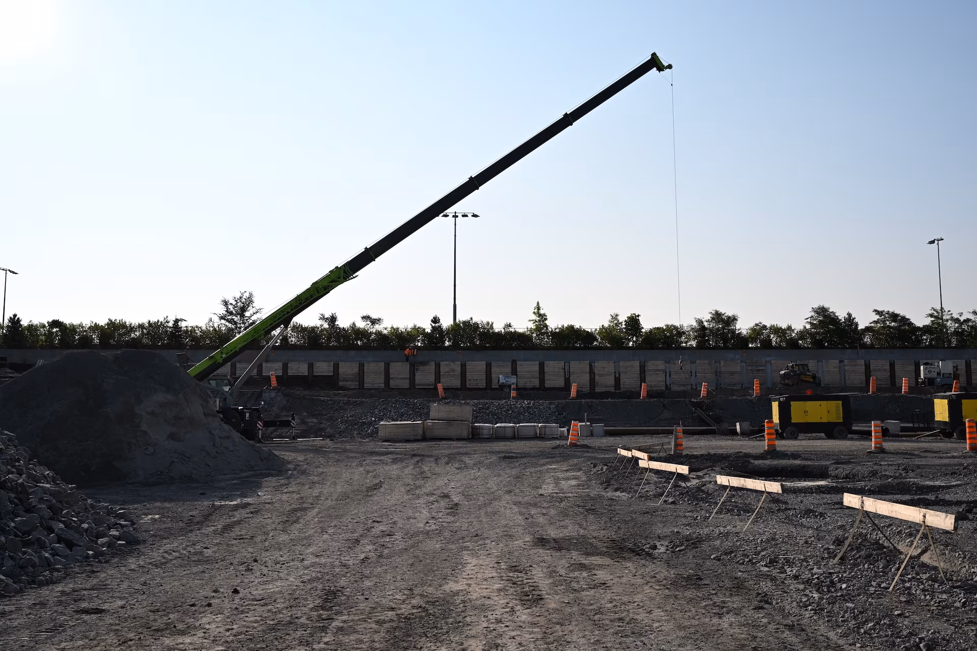 Construction site with a large green crane, piles of gravel, orange safety barrels, and stacked concrete materials under a clear sky.