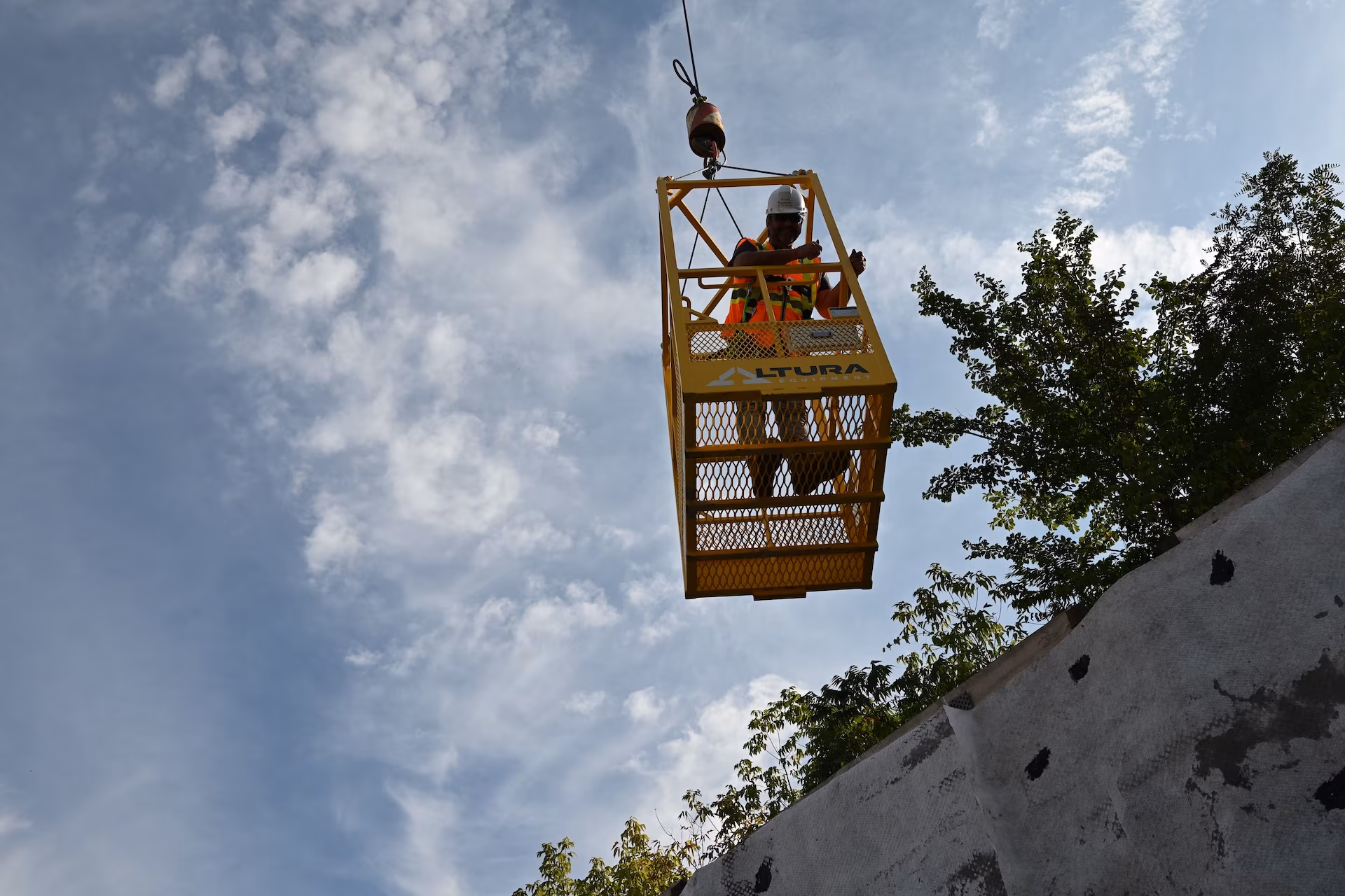 Construction worker wearing safety gear operating a yellow suspended platform against a partly cloudy sky.